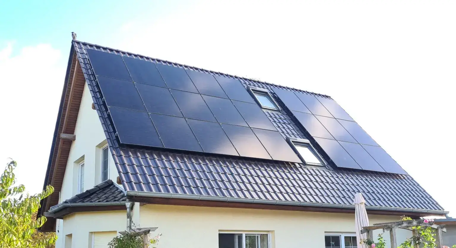 Photovoltaik Berlin 2025: Neue Chancen und Förderungen Einfamilienhaus mit cremefarbenem Putz und steilem, anthrazitfarbenem Pfannendach: Die Sonnenseite ist fast vollflaechig mit schwarzen Modulen belegt, zwei Dachfenster bleiben ausgespart. Rechts eine Pergola mit geschlossenem Sonnenschirm, der Himmel ist hell und diesig.