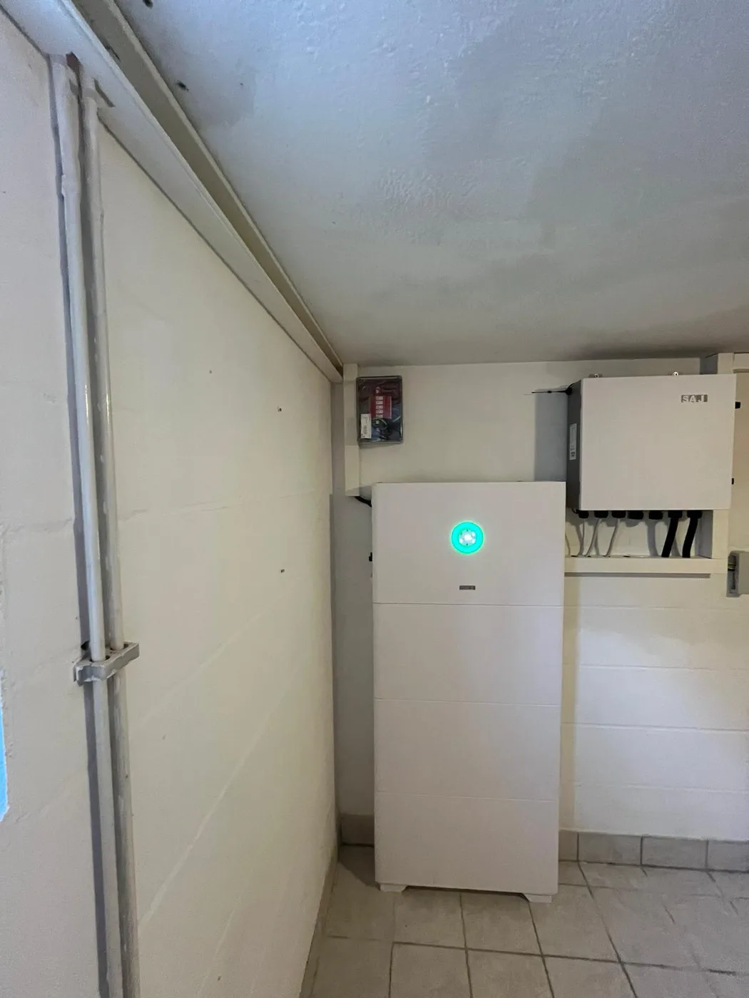 Photovoltaik-Referenz SolarX – Speicher neben dem Verteilerschrank SolarX Referenz: Speicher in einem gefliesten Kellerflur, daneben der geöffnete Verteilerschrank