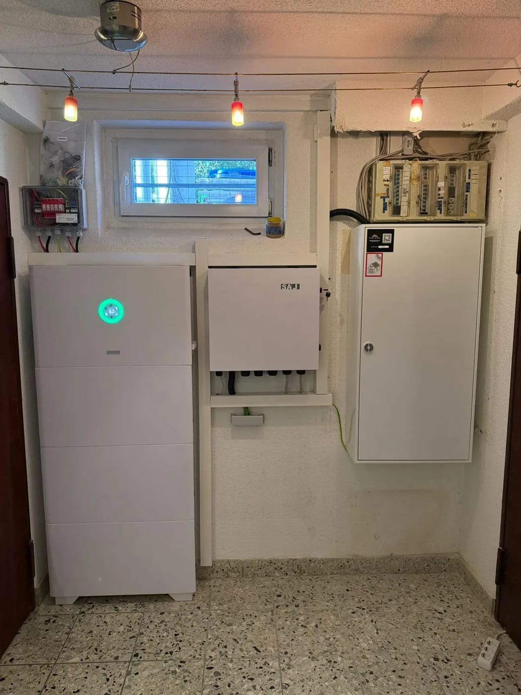 Photovoltaik-Referenz SolarX – Speicher, Wechselrichter und Zählerschrank SolarX Referenz: Speicher, Wechselrichter und Zählerschrank nebeneinander in einem Kellerraum mit Fenster